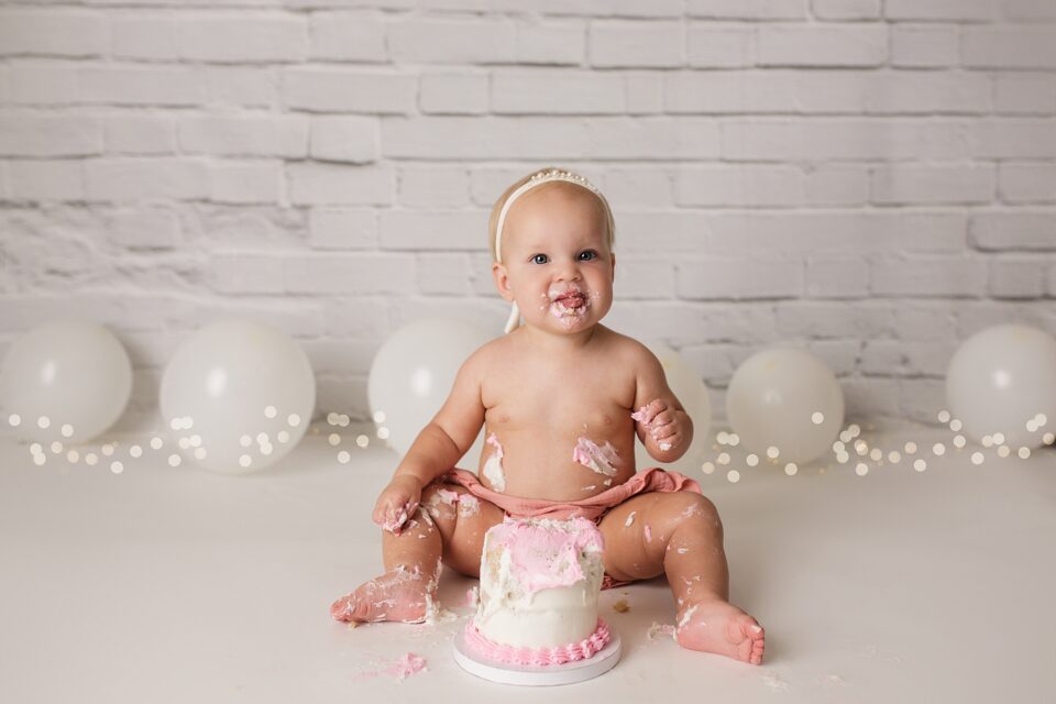 Jackson Missouri newborn photographer, Perryville cake smash photographer, Cape Girardeau newborn photographer, cake smash photoshoot, white brick backdrop, simple cake smash set up, baby eating cake and smiling