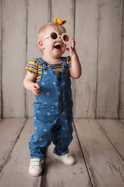 perryville newborn photographer, Cape Girardeau newborn photographer, perryville cake smash photographer, little girl, flower sun glasses, giggle, wood floor, first birthday photoshoot