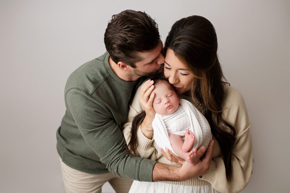 st Louis newborn photographer, farmington Missouri Newborn photographer, cape girardeau Missouri Newborn photographer, perryville, baby boy snuggled by mom, parents kissing baby, white studio backdrop, newborn photoshoot